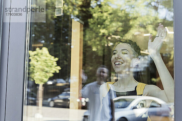 Lächelnde nicht-binäre Person winkt durch Glasfenster