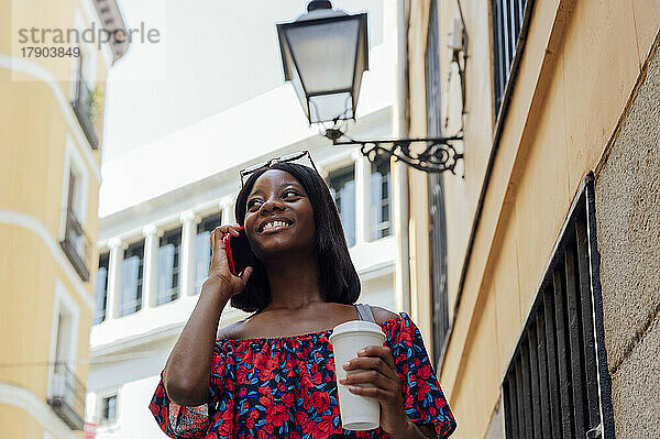 Lächelnde Frau hält eine Einweg-Kaffeetasse in der Hand und telefoniert mit dem Gebäude