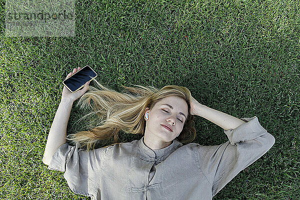 Frau mit geschlossenen Augen hält ihr Mobiltelefon in der Hand und hört Musik über drahtlose In-Ear-Kopfhörer  die im Gras liegen