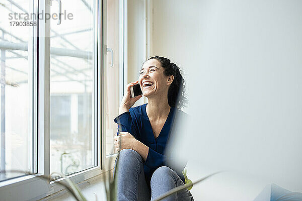 Glückliche Frau  die am Fenster sitzt und mit dem Handy spricht