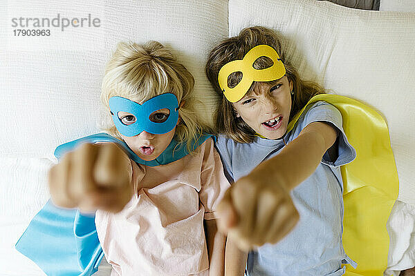 Sisters wearing superhero costume gesturing fists on bed at home