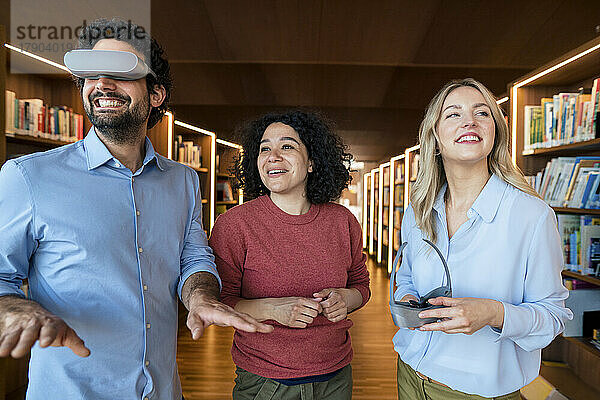 Lächelnder Mann beobachtet Kollegen in der Bibliothek durch eine VR-Brille