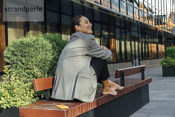Lächelnde Geschäftsfrau entspannt sich auf einer Bank im Büropark