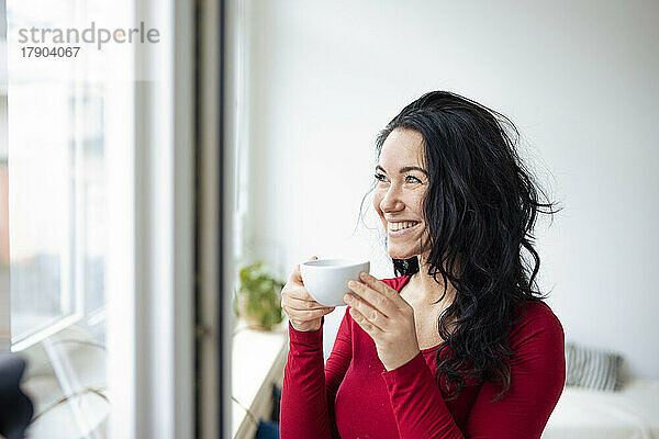 Glückliche Frau mit Kaffeetasse und Blick durch das Fenster