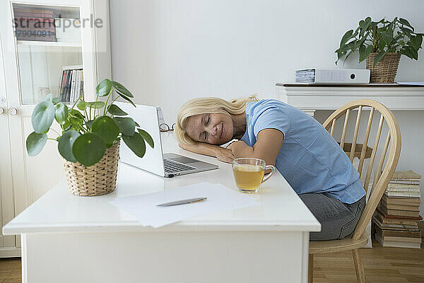 Lächelnder Freiberufler mit geschlossenen Augen entspannt sich am Schreibtisch im Heimbüro