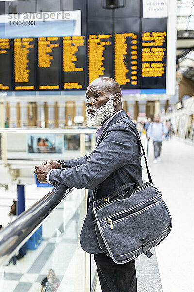 Nachdenklicher Geschäftsmann mit Laptoptasche lehnt am Geländer am Bahnhof
