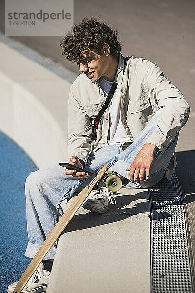 Lächelnder Mann mit Skateboard und Smartphone sitzt am Rand der Leichtathletik