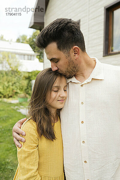 Affectionate father kissing daughter with eyes closed in back yard