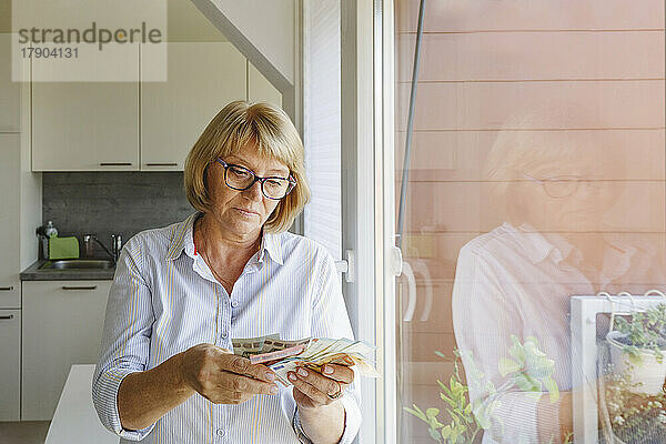 Ältere Frau zählt Papiergeld und steht zu Hause am Glasfenster