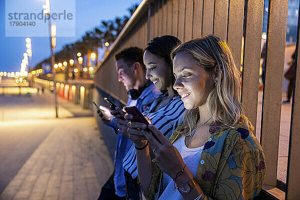 Lächelnde Frauen und Männer  die nachts vor dem Geländer Textnachrichten über Mobiltelefone verschicken