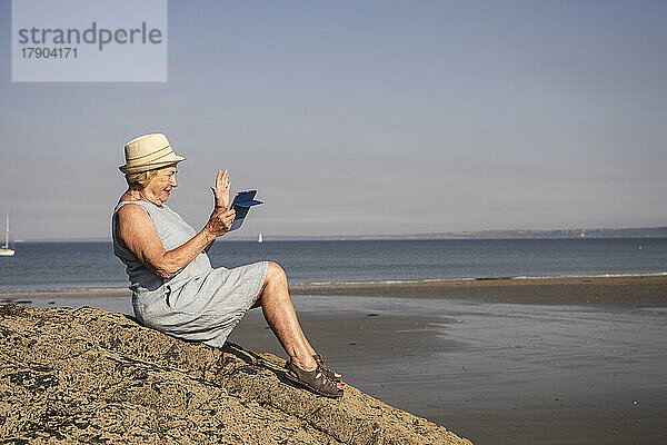 Lächelnde ältere Frau winkt bei einem Videoanruf am Strand