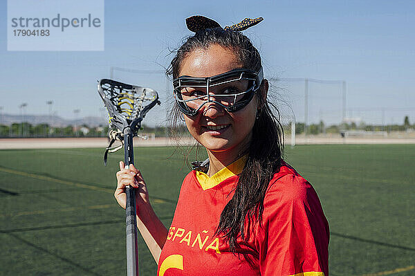 Lächelnder Spieler mit Schutzbrille  der an einem sonnigen Tag einen Lacrosse-Stick hält