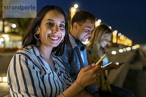 Glückliche junge Frau mit Smartphone  die nachts bei Freunden sitzt