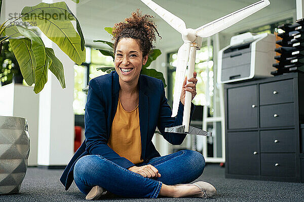 Fröhliche Geschäftsfrau mit Windturbinenmodell sitzt im Büro