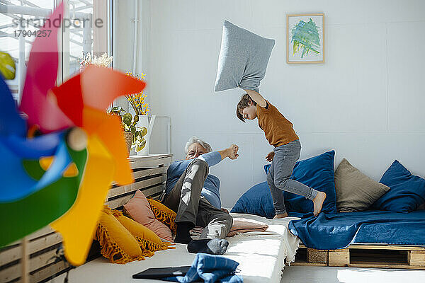 Fröhlicher Junge spielt Kissenschlacht mit Großvater im heimischen Wohnzimmer