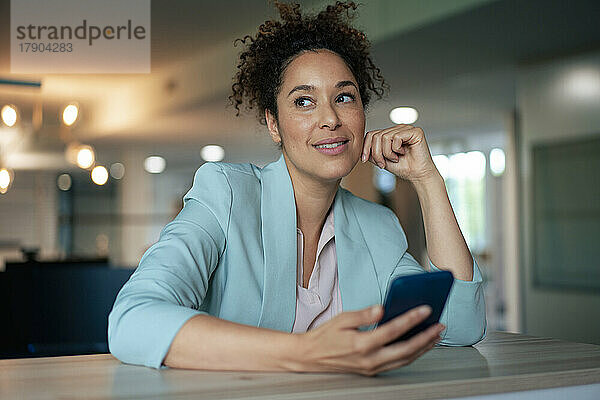 Nachdenkliche Geschäftsfrau mit Mobiltelefon am Tisch im Büro
