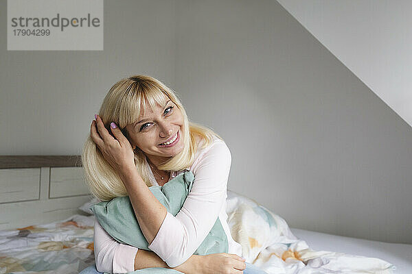 Happy blond woman with pillow sitting on bed at home