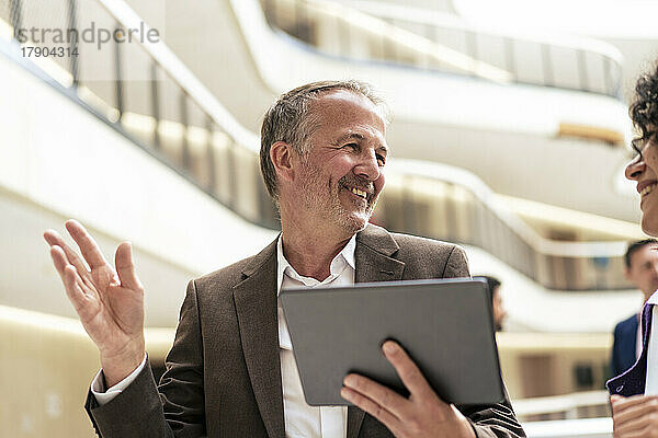 Lächelnder Geschäftsmann hält Tablet-PC in der Hand und diskutiert mit Kollegen im Flur