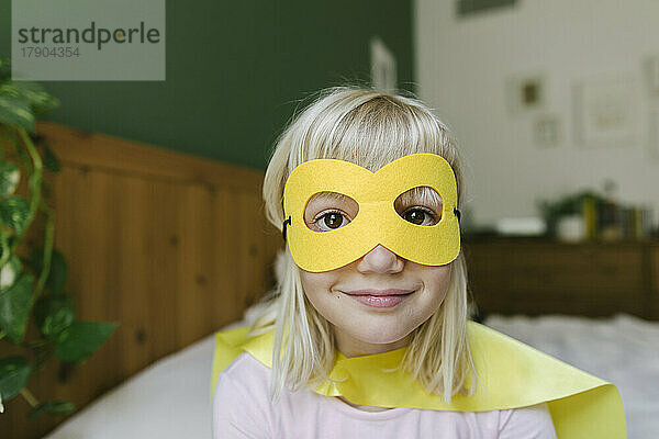 Lächelndes blondes Mädchen mit gelber Maske und Umhang zu Hause