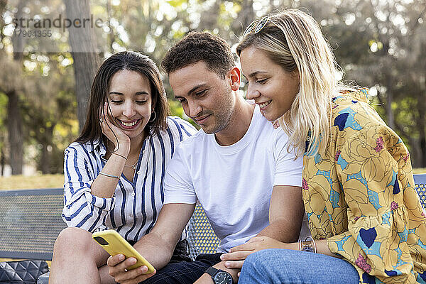 Smiling young man sharing smart phone with friends at park