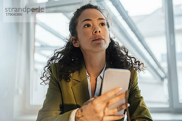 Nachdenkliche Geschäftsfrau mit Smartphone sitzt im Büro