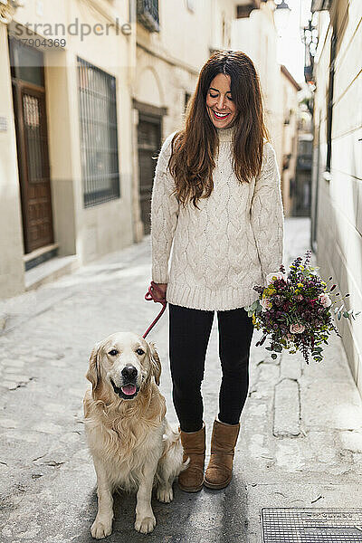 Lächelnde Frau mit Hund hält Blumenstrauß in der Gasse