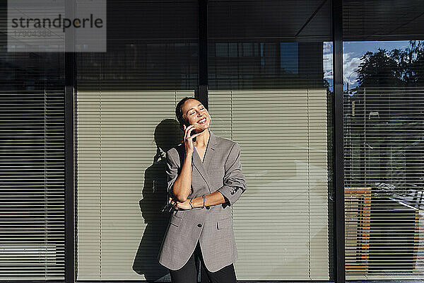 Lächelnde Geschäftsfrau mit geschlossenen Augen  die vor einer Glaswand mit dem Mobiltelefon spricht