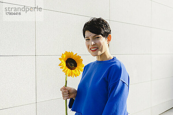 Glückliche Frau mit Sonnenblume  die an einem sonnigen Tag an der Wand steht