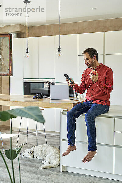 Lächelnder Mann benutzt Smartphone und isst zu Hause Apfel