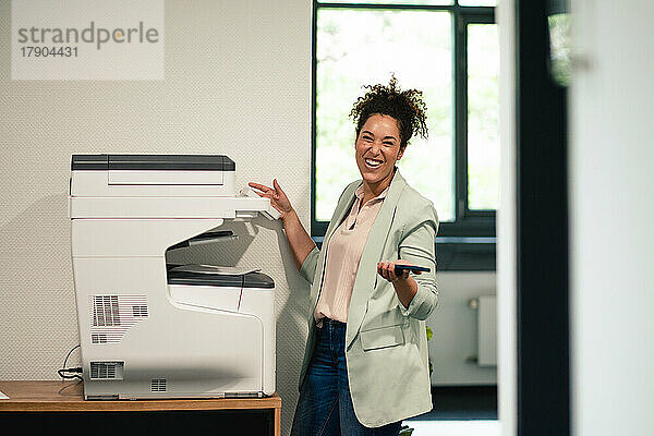 Fröhliche Geschäftsfrau steht am Arbeitsplatz am Computerdrucker
