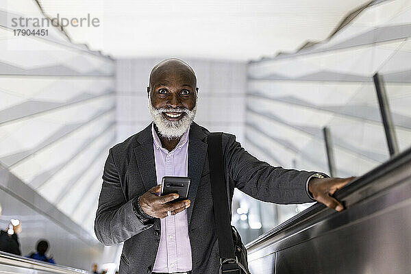 Lächelnder kahlköpfiger Pendler mit Smartphone auf Rolltreppe an U-Bahn-Station