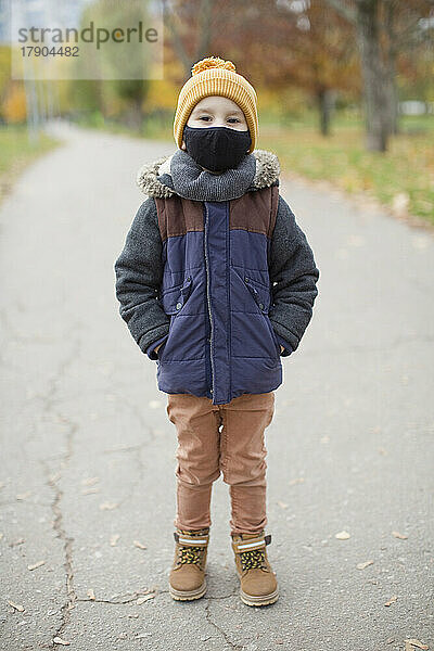 Junge steht mit Händen in den Taschen und trägt eine schützende Gesichtsmaske auf dem Fußweg