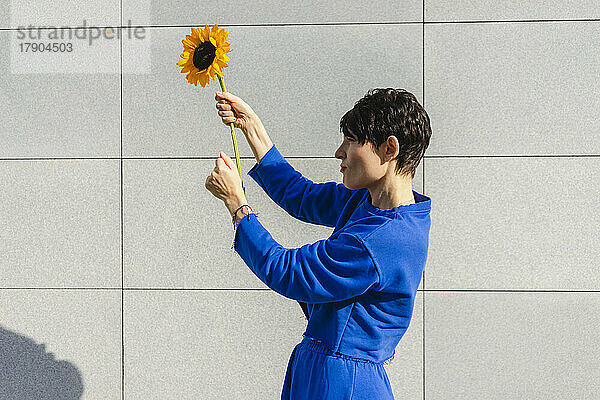 Frau hält an einem sonnigen Tag eine Sonnenblume vor der Wand