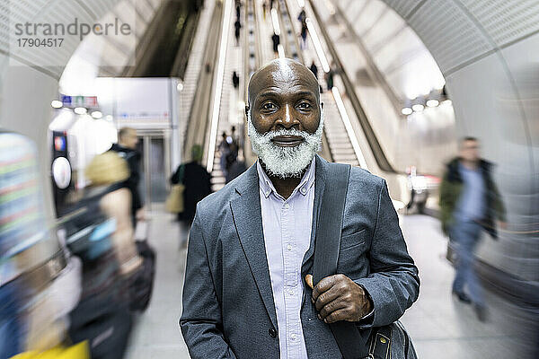 Lächelnder kahlköpfiger Geschäftsmann mit Laptoptasche inmitten von Pendlern an der U-Bahn-Station