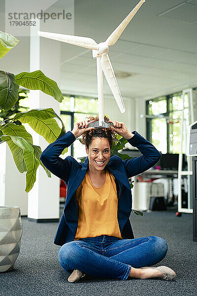 Glückliche Geschäftsfrau trägt Windturbinenmodell auf dem Kopf im Büro