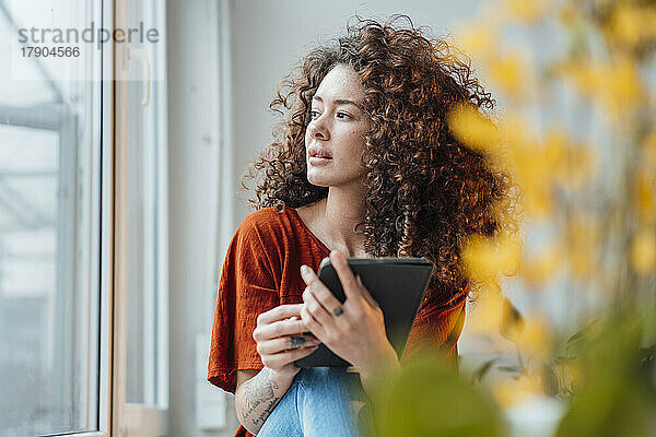 Nachdenkliche Frau mit Tablet-PC sitzt zu Hause am Fenster