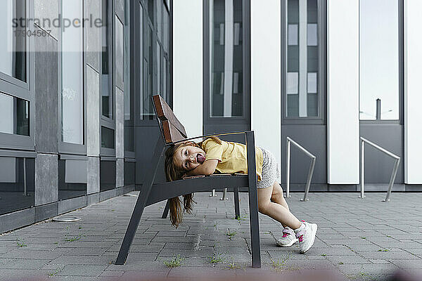 Playful girl sticking out tongue on bench in front of building