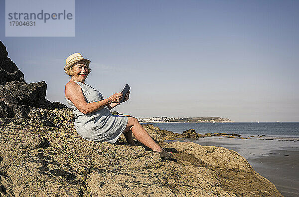 Glückliche Frau mit Tablet-PC sitzt auf einem Felsen am Strand