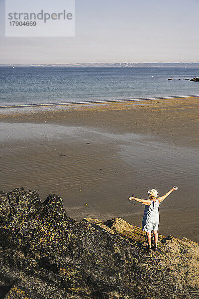 Ältere Frau mit erhobenen Armen steht auf einem Felsen am Strand