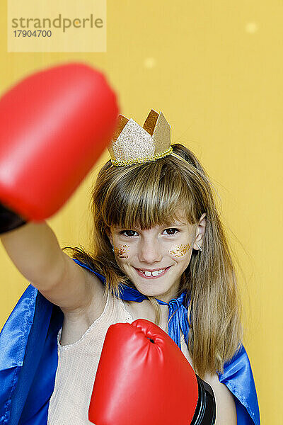 Fröhliches Mädchen mit Boxhandschuhen und Krone vor gelbem Hintergrund