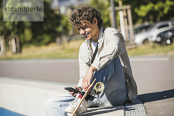 Glücklicher Mann mit Skateboard und Smartphone am Streckenrand sitzend