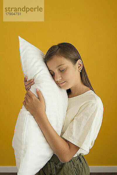 Girl with eyes closed holding white pillow in front of yellow wall