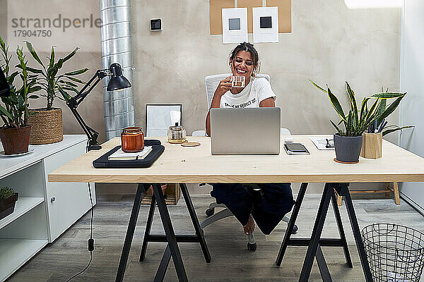 Lächelnde Geschäftsfrau trinkt Tee und macht Videoanrufe über Laptop im Heimbüro