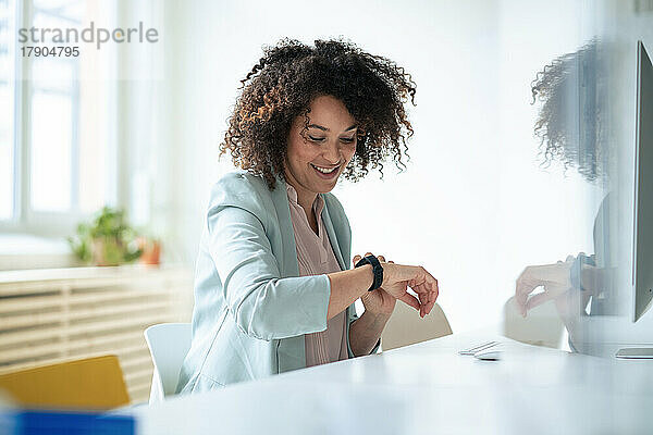 Lächelnde Geschäftsfrau mit lockigem Haar  die im Büro die Zeit auf einer Smartwatch überprüft