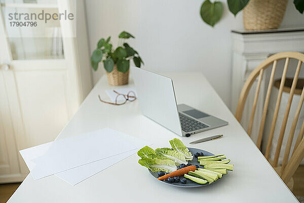 Gemüse auf dem Teller und Laptop auf dem Schreibtisch im Heimbüro