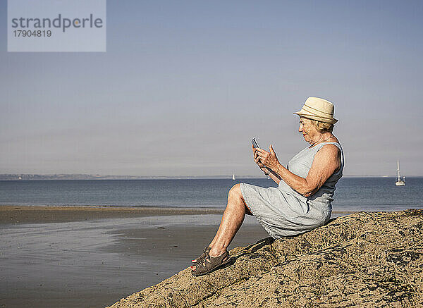 Ältere Frau sitzt mit Tablet-PC auf einem Felsen am Strand