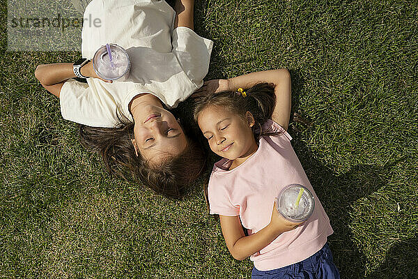 Smiling girls with milkshakes lying together on grass at park