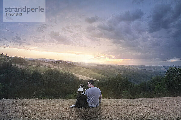 Ein Mann und sein Border Collie Hund sitzen auf einem Feld in der Landschaft und genießen den Sonnenuntergang  Italien  Europa