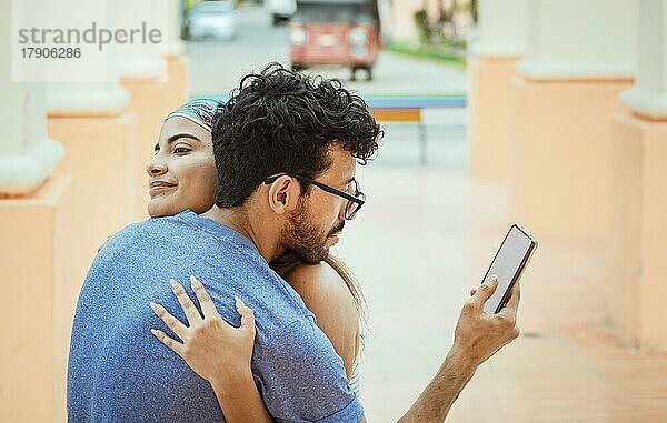 Untreuer Mann schaut auf das Handy  während er seine Freundin im Freien umarmt. Untreuer Freund umarmt seine Freundin und Blick auf das Handy. Konzept der unfaithful Mann mit Handy