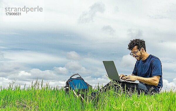 Lächelnder Mann mit Laptop sitzt auf dem Gras im Feld. Seitenansicht des Mannes sitzt auf grünem Gras arbeiten mit seinem Laptop. Lächelnd Freiberufler Kerl arbeiten aus dem Feld